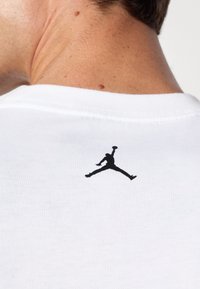 White cotton t-shirt featuring a black Jumpman logo printed on the back near the neck. Short sleeves, crew neckline, smooth texture.