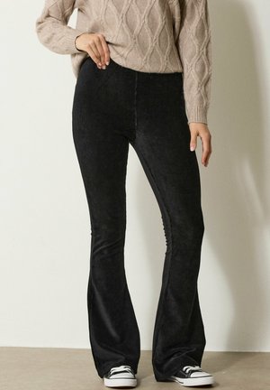 Femme portant un pantalon en velours côtelé noir à jambes évasées, un pull en mailles beige, et des baskets noir et blanc, se tenant contre un mur uni.