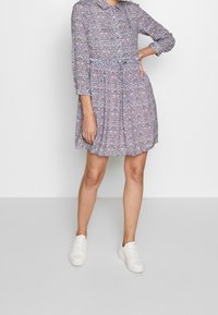 Blumenmuster-Kleid aus Stoff mit Kragen, langen Ärmeln, tailliertem Oberteil und ausgestelltem Rock. Kombiniert mit weißen Sneakers.