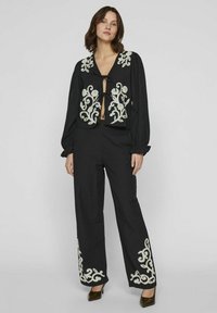Conjunto de dos piezas negro que incluye un top corto con bordado floral blanco y mangas largas, combinado con pantalones de pierna ancha y tacones.