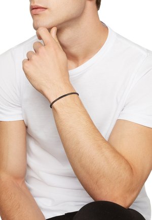 Zwarte leren armband om de pols, gladde textuur, minimalistisch ontwerp, gedragen over een wit T-shirt; arm rustend nabij de kin.