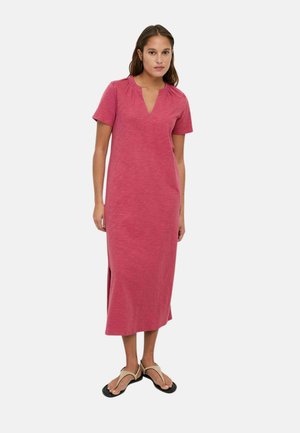 Mujer de pie que lleva un vestido midi de manga corta de color rosa con aperturas laterales y zapatos estilo sandalia beige sobre un fondo liso.