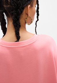 Rosa genser med rund hals og teksturert stoff. Et gullfarget, bølgete hoop ørepynt er synlig på modellen. To fletter er stylet bak.