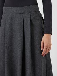 Mujer vistiendo una falda plisada gris oscuro y una camiseta de manga larga negra, con la mano relajada a un lado, contra un fondo claro y liso.