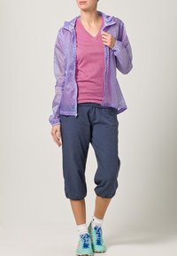 Světle fialová vzorovaná bunda s kapucí, nošená přes růžové tričko s výstřihem do V a tmavě modré zkrácené kalhoty. Doplněno vícebarevnými sportovními botami.