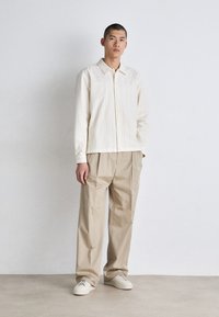 Camicia col beige chiaro con colletto e righe verticali, abbinata a pantaloni beige larghi e sneakers color crema; texture liscia e vestibilità rilassata.