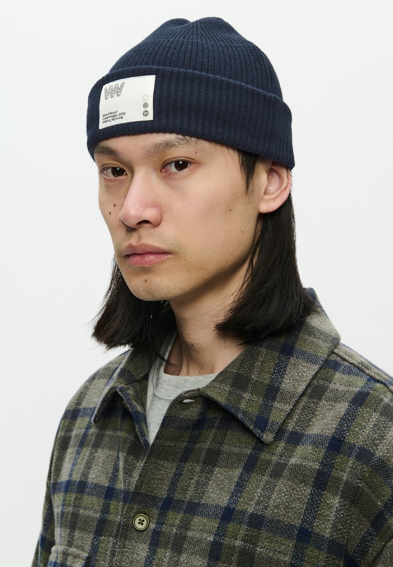 Young person with long dark hair wearing a navy knit beanie with a white patch, plaid jacket in green and blue tones, and a light gray shirt.