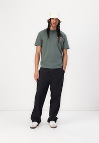 Green cotton t-shirt with a printed design, paired with loose-fitting black pants and white sneakers. Bucket hat in light beige color.