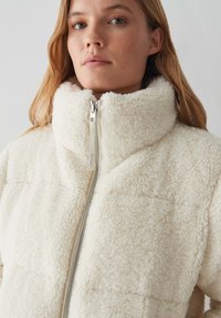 Giacca in pile bianco con colletto alto, dotata di chiusura con zip e superficie morbida e con texture. Il design è trapuntato con cuciture evidenti.