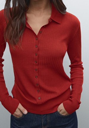 Femme portant un pull rouge ajusté à manches longues en maille côtelée avec boutons et col, mains dans les poches, et un jean bleu foncé.