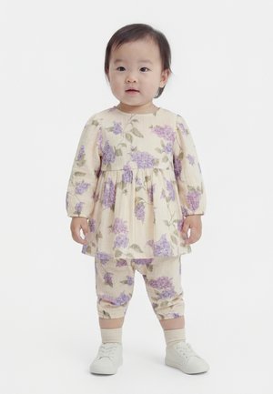 Småbarn stående i et cremefarvet outfit med lilla blomsterprint, lange ærmer, bukser med kort ben, hvide sko og lyse sokker på en hvid baggrund.