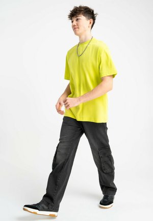Młoda osoba spacerująca, ubrana w jaskrawożółtą koszulkę, czarne spodnie cargo, czarne sneakersy oraz srebrny łańcuszek, na białym tle.