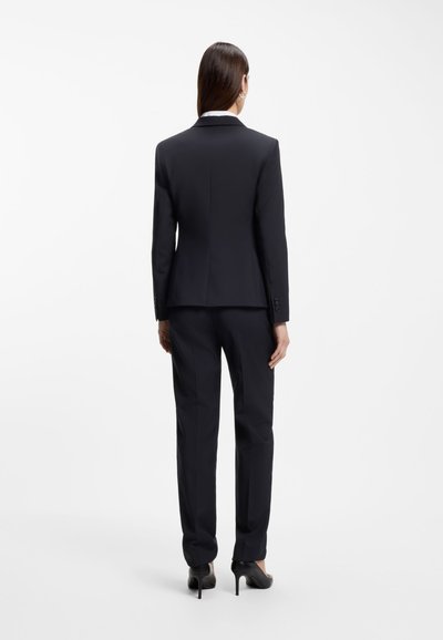 Femme debout, de dos, portant un costume noir sur mesure avec un pantalon droit et des talons hauts noirs sur un fond blanc.