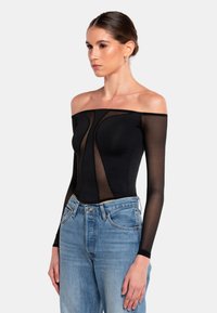 Schwarzer Off-Shoulder-Bodysuit mit transparenten langen Ärmeln, figurbetonter Passform und geschwungenen Paneeldetails. Kombiniert mit hellblauen Jeans.