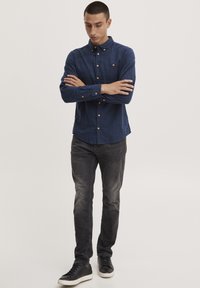 Mørkeblå skjorte med krage, med gullknapper og brystlomme, sammen med mørkegrå jeans og svarte joggesko.