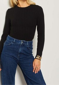 Haut noir à manches longues côtelé associé à un jean denim bleu taille haute. Le poignet du modèle est orné de bracelets dorés. Tissu texturé visible.