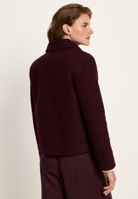 Woman with short brown hair wearing a dark burgundy wool jacket and matching pants, standing against a plain light background.