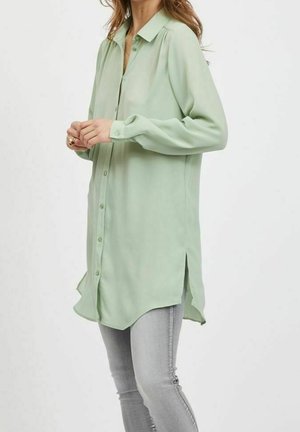 Vrouw draagt een losse, lichtgroene blouse met lange mouwen en knopen, voorzien van zij-splitten, gecombineerd met nauwsluitende lichtgrijze jeans.