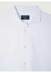 Witte poloshirt met een ribbelstructuur, drie knopen en een klein geborduurd logo aan de linkerkant. Merklabel zichtbaar onder de kraag.