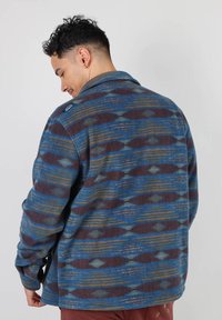 Veste en mélange de laine présentant une base bleue avec des motifs marron et jaune, avec un col et des manches longues, vue de dos.