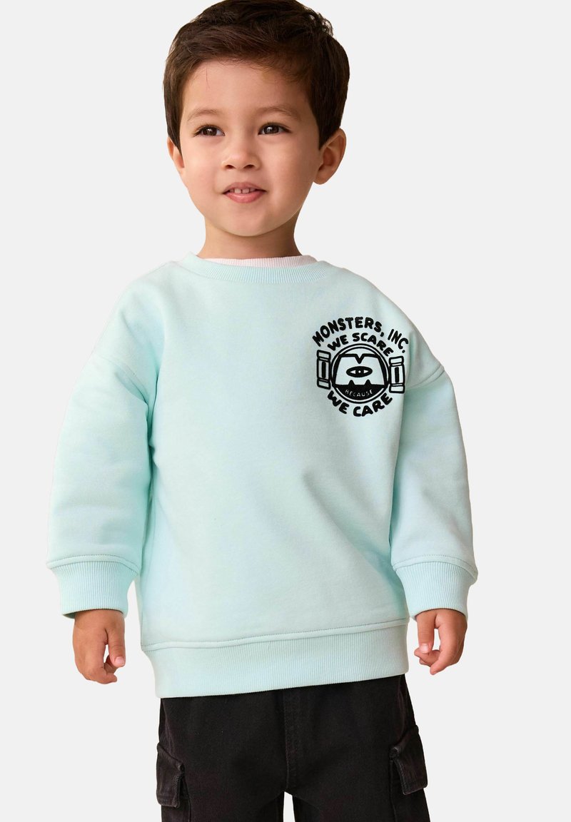 Jeune garçon portant un sweat-shirt bleu clair avec un logo Monsters, Inc. et un pantalon noir sur un fond clair uni.