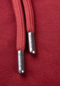 Rote geflochtene Kapuzenzugbänder mit metallischen Enden, die "SPITZBUB" eingraviert haben, auf einem strukturierten Stoffhintergrund.