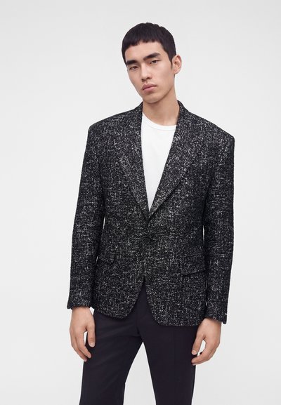 Jeune homme aux cheveux courts et noirs portant un blazer texturé noir et blanc par-dessus une chemise blanche et un pantalon noir sur un fond uni.