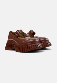 Chaussures Mary Jane synthétiques marron avec deux sangles à boucle et une semelle épaisse à rainures. Présente des détails en relief sur la pointe.