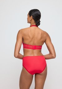 Modelka stoi tyłem, ubrana w jasnoczerwony stanik bikini i wysokie majtki na tle jasnego tła.