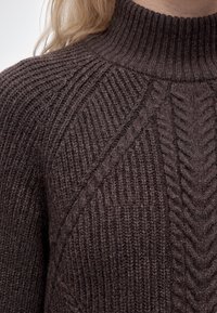 Gros plan d'un pull tricoté marron foncé avec un col montant côtelé et des motifs torsadés tressés sur l'épaule et la poitrine.