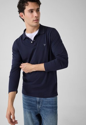 Jeune homme remontant les manches d'un polo bleu marine à manches longues avec un petit logo, portant un jean bleu clair sur un fond uni.