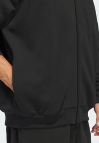 Crna majica s patentnim zatvaračem izrađena od mekanog materijala. Ima rebraste manšete i rub te bočni džep. Jednostavan dizajn, bez vidljivih uzoraka ili grafika.