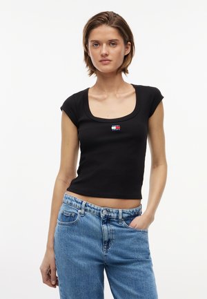 Jeune femme aux cheveux courts et bruns, portant un haut noir ajusté Tommy Jeans et un jean taille haute bleu clair, debout avec une main dans la poche.