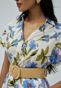 Vestido tipo camisa con estampado floral que presenta flores azules sobre un fondo crema, mangas cortas, cierre de botones y un amplio cinturón de fibra natural con una hebilla redonda.