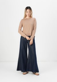 Beige lange mouwen trui met een ontspannen pasvorm en ronde hals, gecombineerd met wijde donkere denimbroek, staande voor een effen achtergrond.