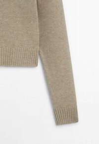 Manche et bas du torse d'un pull beige tricoté avec des poignets et un ourlet côtelés sur un fond blanc.