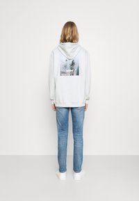 Ljust grå luvtröja med en stor grafisk av palmträd på baksidan. Tillsammans med blå jeans och vita sneakers ger outfiten ett avslappnat intryck.