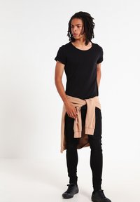 Zwarte T-shirt met opgerolde korte mouwen, gecombineerd met aansluitende zwarte broek. Een lichtbeige sweatshirt is om de taille geknoopt. Zwarte schoenen.