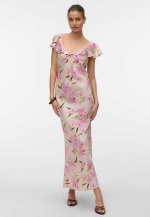 Kvinne står i en lang rosa blomstret kjole med flagrende ermer, iført brune sandaler og et armbånd på venstre håndledd.