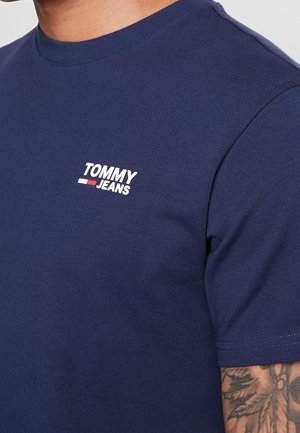 Close-up of a person wearing a navy blue Tommy Jeans t-shirt with a tattooed arm visible at the sleeve.