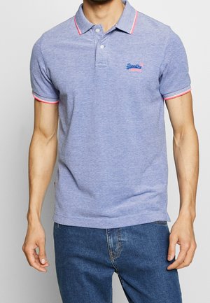 Homme portant un polo bleu clair avec des bords roses sur le col et les manches, assorti à un jean bleu, debout devant un fond uni.