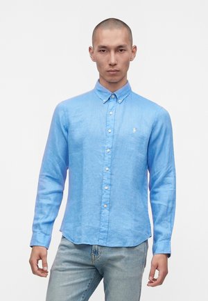 Homme aux cheveux coupés courts portant une chemise bleu clair à boutons et un jean délavé clair, debout devant un fond blanc uni.
