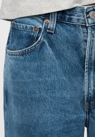 Κοντινή λήψη της μέσης και της μπροστινής τσέπης μπλε τζιν με μεταλλικά καρφιά και κουμπί, φορεμένο με ανοιχτό γκρι τοπ.