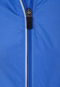 Blå jacktyg med en slät textur, som har en central dragkedja i kontrasterande blått, samt en svart dragkedjeh e med en logotyp.