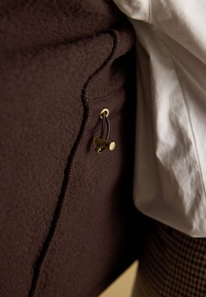 Nærbillede af mørkebrun fleecejakke med gyldent spænde og snøre lukning ved siden af hvidt stof og mønstrede bukser.