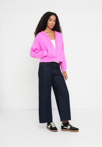 Felpa cropped fucsia con tasche anteriori, abbinata a jeans in denim scuri a gamba ampia e sneakers nere con accenti bianchi e suole in gomma.