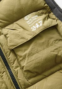 Veste matelassée kaki avec fermeture éclair, grande poche avant et logo imprimé avec motif de globe, contrastée par des finitions foncées.