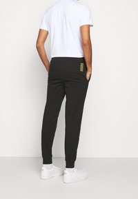 Svarta joggerbyxor med en smalnande design, med en subtil gyllene randdetalj på bakfickan och elastiska muddar. Bärs med en vit skjorta.