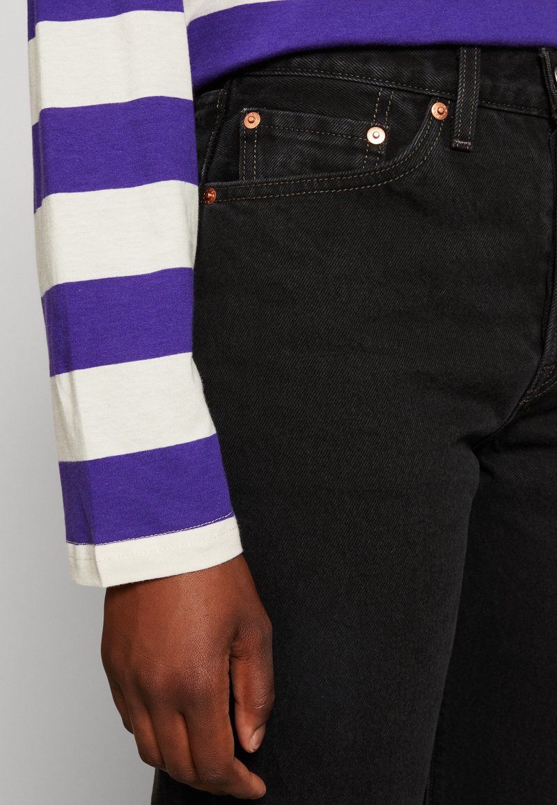 Jeans de mezclilla negra con remaches y costuras de cobre, combinados con una camisa de manga larga a rayas crema y púrpura. Vista cercana de la mano y la ropa.