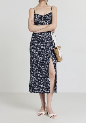 Kvinde iført en sort og hvid blomsterkjole med spaghetti-stropper og en høj slids på låret, holder en flettet håndtaske og har hvide sandaler med hæl på.